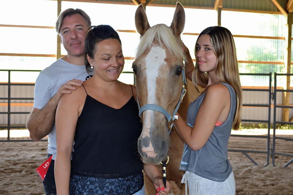 A father and mother stand on the left of a horse, their teenage daughter stands on the right. All seem happy and content.